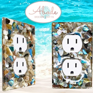 May include: Decorative electrical outlet covers with a mosaic of colorful shells and stones. The covers feature white outlets and are set against a beach-themed background. The Abode Decor Gifts logo is visible.
