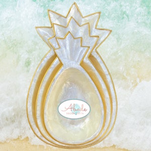 May include: A set of three white and gold pineapple-shaped bowls. The bowls are made of mother of pearl and have a gold rim. The bowls are nested inside each other.