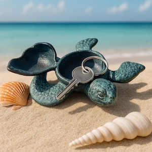 May include: A teal-colored turtle-shaped key holder with a key resting inside. The key holder is on a sandy surface with seashells and the ocean in the background. The key holder is made of metal and has a textured surface.