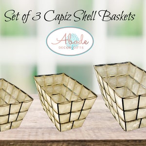 May include: Set of three capiz shell baskets in different sizes. The baskets are made of white capiz shells and have a gold trim. The baskets are arranged in a row on a wooden surface.