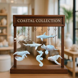 May include: A wooden display stand with the text "COASTAL COLLECTION" features hanging sea creature ornaments. The ornaments include a crab, sharks, a seahorse, a turtle, and a whale, all in shades of white and blue. The stand is brown.