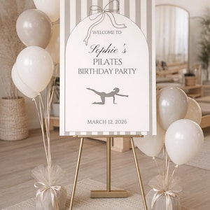 Puede incluir: Un caballete dorado exhibe un letrero para la fiesta de cumpleaños de Pilates de Sophie, con globos blancos y beige. El letrero tiene un lazo gráfico y la fecha 12 de marzo de 2026.