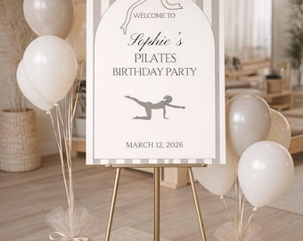 May include: A gold easel displays a sign for Sophie's Pilates Birthday Party, with balloons in white and beige. The sign has a bow graphic and the date March 12, 2026.