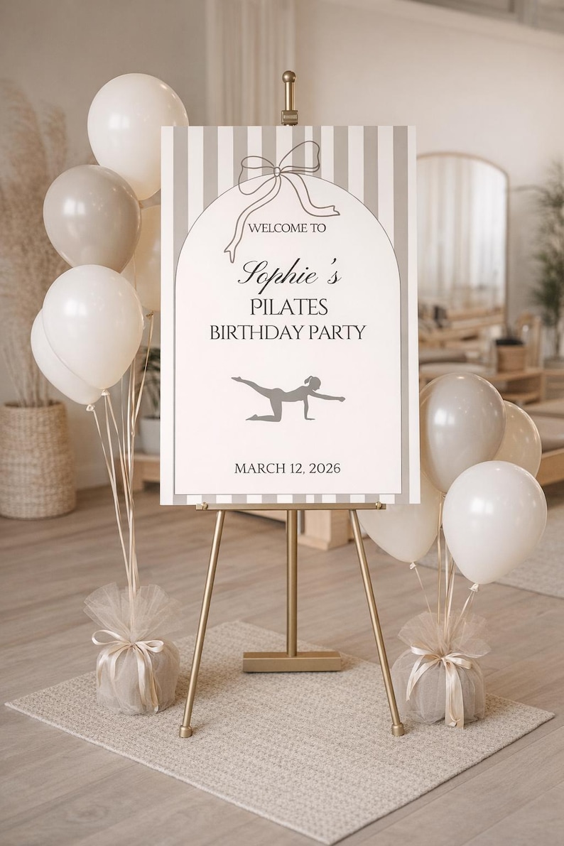 Peut inclure: Un chevalet dor&eacute; pr&eacute;sente une affiche pour l'anniversaire Pilates de Sophie, avec des ballons blancs et beiges. L'affiche comporte un n&oelig;ud graphique et la date du 12 mars 2026.