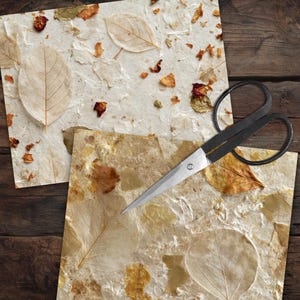 May include: Two sheets of textured, off-white handmade paper embedded with dried leaves and flower petals, featuring a visible vein structure on the leaves. A pair of black metal scissors with large finger loops rests on the paper sheets.