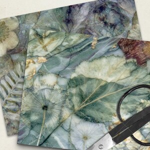 May include: Close-up of two decorative paper sheets with a botanical design. The sheets feature a marble-like pattern in shades of green, blue, and gold, with leaf and flower imprints. A pair of scissors is in the foreground.