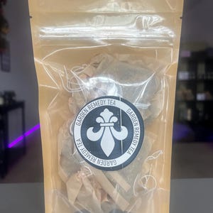 May include: A transparent plastic bag containing tea bags, sealed with a brown paper backing. The bag features a circular label with the text "GARDEN REMEDY TEA" and a white fleur-de-lis design.