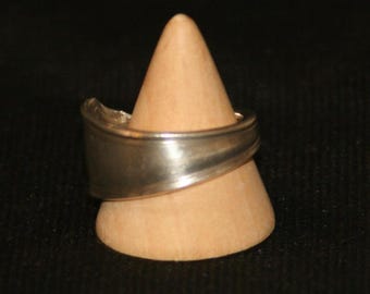 Könnte beinhalten: Ein silberner Ring aus einem Löffel, präsentiert auf einem Holzkegel. Der Ring hat ein geschwungenes Design und eine polierte Oberfläche, die das recycelte Besteck hervorhebt. Ein einzigartiges Schmuckstück.