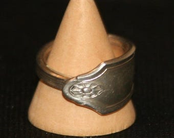 Könnte beinhalten: Ein handgefertigter Silberring aus einem Löffel, mit einem dekorativen Blumendesign auf der Löffelschale. Der Ring wird auf einem hellbraunen, kegelförmigen Ständer vor dunklem Hintergrund präsentiert. Das Band des Rings ist aus dem Löffelstiel geformt.