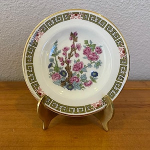 May include: A small white ceramic plate with a floral design in shades of pink, green, and blue. The plate has a gold rim and a green and gold border with a repeating geometric pattern. The plate is on a gold metal stand.