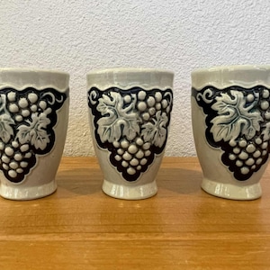 Puede incluir: Tres vasos de cerámica con base color crema y diseño de uvas azul oscuro. El diseño de uvas incluye hojas y un racimo de uvas. Los vasos están sobre una superficie de madera.