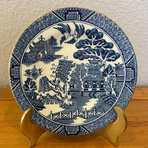 Puede incluir: Un platillo de porcelana con diseño de sauce azul y blanco con borde dorado. El platillo está sobre un soporte de metal dorado.