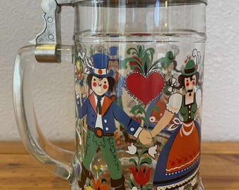 May include: Clear glass beer stein with a hinged metal lid and handle. The stein is decorated with a colorful illustration of a couple in traditional clothing, holding hands. Floral and heart motifs are also present.