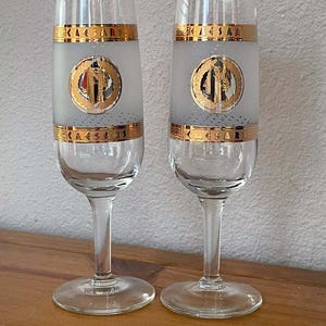 May include: Two clear glass champagne flutes featuring a frosted middle section with a gold-rimmed band at the top and bottom. Each glass displays a circular gold emblem with a central graphic and the text CAESARS PALACE LAS VEGAS.