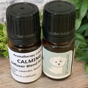Könnte beinhalten: Zwei kleine braune Glasflaschen mit schwarzen Deckeln, beschriftet mit "CALMING Diffuser Blend for Pets" und einer Liste ätherischer Öle: Weihrauch, Lavendel und römische Kamille. Das Etikett weist auch darauf hin, 2 Tropfen in einen Diffusor zu geben. Eine Flasche hat ein Cartoon-Bild eines weißen Hundes auf dem Etikett.