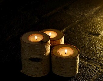 May include: Three small, round, wooden candle holders with a natural birch bark finish. Each holder has a small, white tea light candle inside, and the flames are lit.