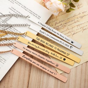 May include: Several personalised metal bookmarks with chain detailing, displayed on an open book. Each bookmark has engraved text and decorative charms. The bookmarks are in silver, gold, and rose gold hues, with different fonts and designs.