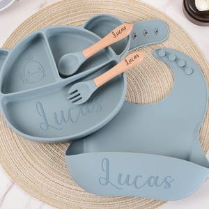 May include: A baby feeding set in a soft blue color, including a divided plate, bib, spoon, and fork. The plate has a whale design and the name "Lucas" is written on the plate and bib. The utensils have wooden handles.