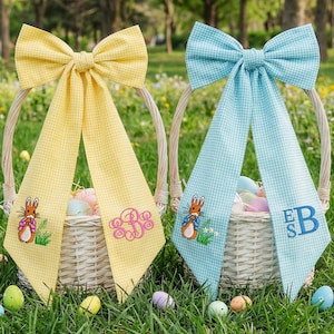 May include: Two Easter baskets with gingham bows, one yellow and one blue, filled with colourful eggs. Each bow has a monogram and a bunny embroidery. The baskets sit on green grass with Easter decorations.