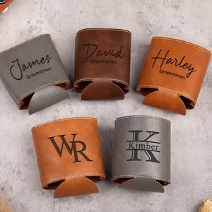 Pode incluir: Cinco mangas cilíndricas para bebidas feitas de material liso em tons de cinza, marrom e bege. Cada manga apresenta texto ou monogramas pretos personalizados, incluindo os nomes James, David, Harley, as iniciais WR e o nome Kimber.