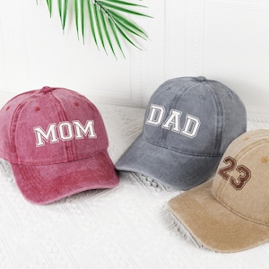 以下が含まれることがあります： スタジオで撮影された3つの野球帽。1つは白のブロック体で「MOM」と書かれた色あせた赤色、白で「DAD」と書かれたグレーの帽子、茶色の数字「23」が付いたタン色の帽子。すべての帽子にはカーブしたつばが付いています。