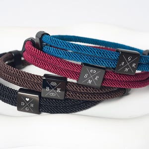 May include: Four braided rope bracelets in black, brown, burgundy, and teal. Each bracelet has a black square clasp with engraved initials and symbols. The bracelets are displayed on a white surface.