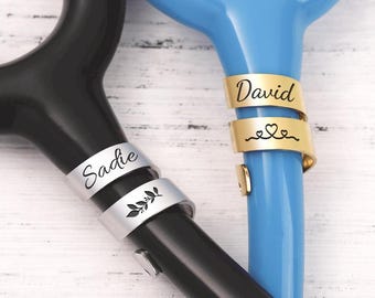 May include: A black stethoscope and a blue stethoscope, each featuring a personalised metal ID tag wrapped around the tubing. The silver tag on the black stethoscope is engraved with the name Sadie and a leaf icon, while the gold tag on the blue stethoscope displays the name David and a heart icon.