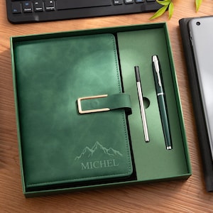 May include: A dark green notebook with a gold-toned rectangular clasp, featuring an engraved mountain graphic and the name MICHEL on the cover. The notebook is presented in a matching green gift box alongside a dark green pen and a metal ink refill.