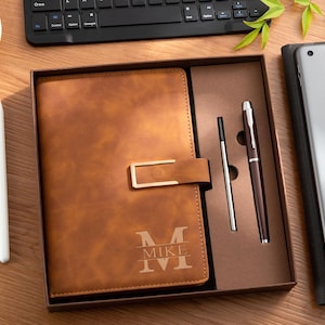 May include: A tan leather-effect notebook with a gold-toned metal clasp and a personalised monogram featuring the name MIKE, presented in a gift box alongside a matching brown ballpoint pen and a single ink refill cartridge.