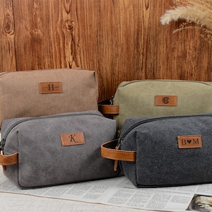 May include: Four canvas toiletry bags in shades of brown, green, and gray, each with a leather handle and a leather patch with initials. The bags are zippered and sit on a newspaper. The background is a wooden wall.
