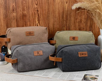 May include: Four canvas wash bags in brown, green, and grey, each with a leather handle and a leather patch bearing initials. The bags have zippers and are on a newspaper. The background is a wooden wall.