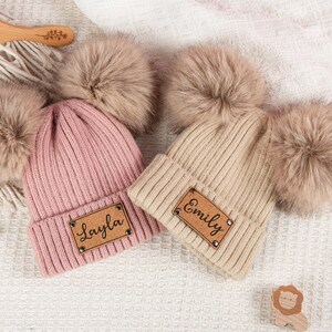 Op de afbeelding: Twee gebreide mutsen met imitatiebont pompons. Eén is roze met een leren label met de naam "Layla", de andere is beige met een label met de naam "Emily". Een houten kam en speelgoed staan ook op de foto.