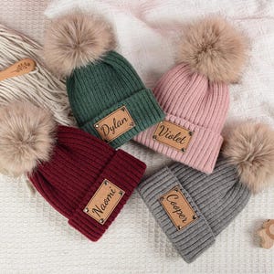 Könnte beinhalten: Vier gestrickte Mützen in Grün, Rosa, Burgunderrot und Grau, jede mit einem Kunstfell-Bommel und einem Namensschild aus Kunstleder. Namen auf den Etiketten sind Dylan, Violet, Naomi und Cooper. Holzspielzeug und eine Holzbürste sind ebenfalls sichtbar.