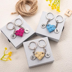 Può includere: Portachiavi con ciondoli a forma di cuore rosa, blu e giallo, ciascuno con un ciondolo con iniziale argentato. I portachiavi sono attaccati ad anelli portachiavi argentati e presentati su piccole scatole regalo argentate. Una corda e lettere di legno sono sullo sfondo.