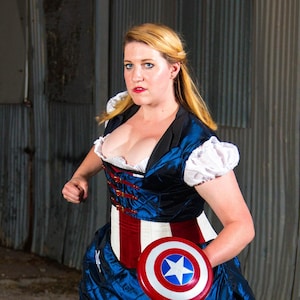 May include: A woman dressed in a blue and red costume with a white star on the belt, holding a red, white, and blue shield with a white star in the center. She is standing in front of a corrugated metal wall.