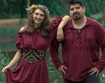 May include: A couple wearing matching burgundy outfits pose in a forest setting. The woman wears a flower crown and a patterned belt. The man has a beard and wears a long-sleeved shirt with a lace-up neckline.