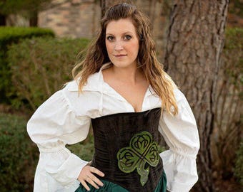 May include: A woman wearing a white blouse with a brown corset featuring a green shamrock design. She is also wearing a green skirt with ruffles.