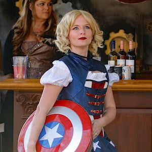 May include: A woman wearing a blue and red vest with a white shirt and a blue and white skirt holds a red, white and blue Captain America shield.