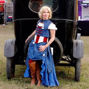 Puede incluir: Una mujer con un cors&eacute; azul y rojo con estrellas blancas y una falda azul con estrellas blancas, de pie junto a un coche negro vintage.