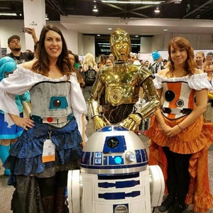 May include: Two people dressed in Star Wars costumes pose with a R2-D2 robot. One person is dressed as C-3PO, a gold robot with a silver chest plate. The other person is dressed as a character from the Star Wars universe, wearing a white and orange costume with a black belt. The robot is white and blue with a round dome.