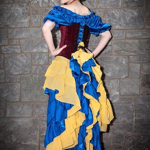 May include: A woman dressed as Snow White in a blue and yellow ruffled skirt, a burgundy corset with a lace-up front, and a blue off-the-shoulder top. She is wearing a red flower in her hair.