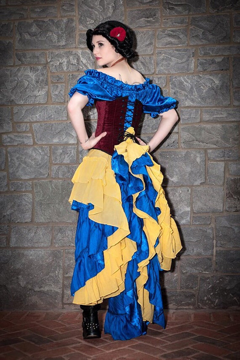 May include: A woman dressed as Snow White in a blue and yellow ruffled skirt, a burgundy corset with a lace-up front, and a blue off-the-shoulder top. She is wearing a red flower in her hair.