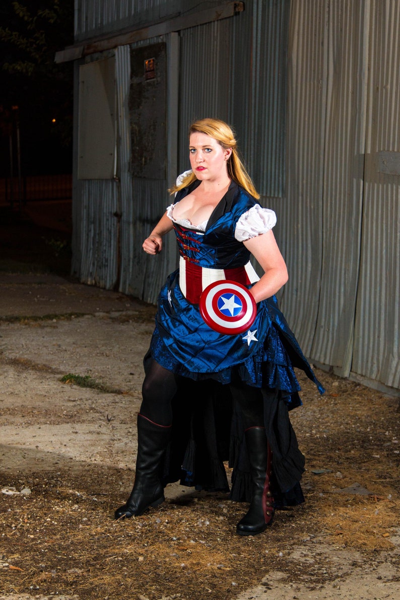May include: A woman dressed in a blue and red costume with a white star on the belt, holding a red, white, and blue shield with a white star in the center. She is standing in front of a corrugated metal wall.