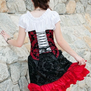 May include: A woman wearing a white blouse, a red and black corset with black lacing, and a black and red skirt with ruffles. The corset has a floral pattern.