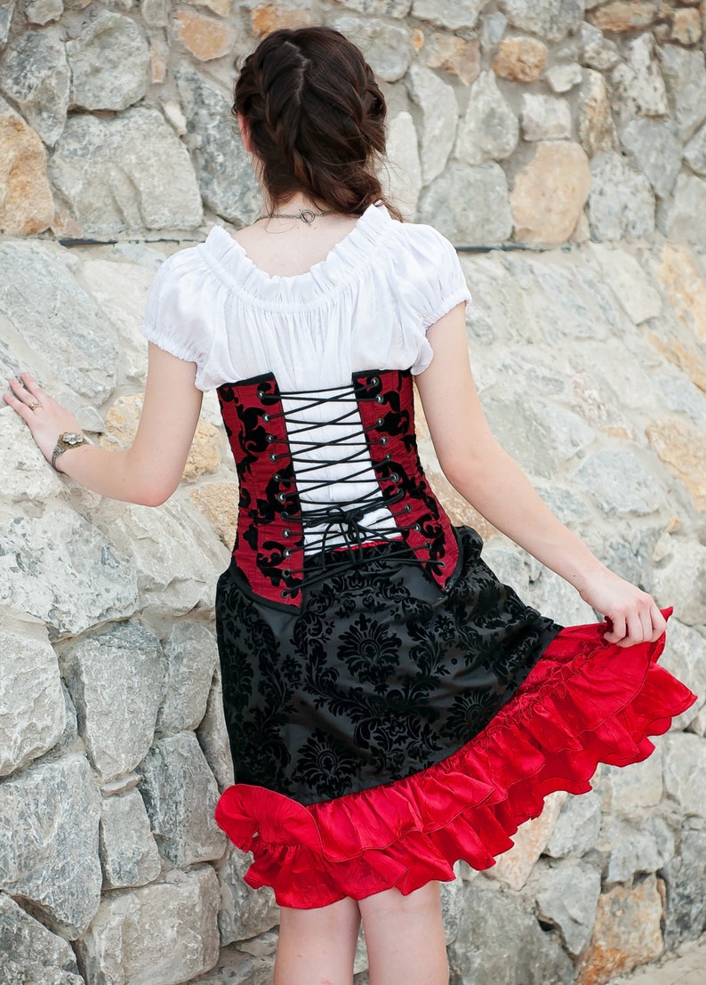 May include: A woman wearing a white blouse, a red and black corset with black lacing, and a black and red skirt with ruffles. The corset has a floral pattern.