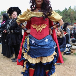 Puede incluir: Una mujer vestida como Wonder Woman con un traje rojo y azul con una falda de estrellas doradas y una capa roja. Lleva una corona dorada con una estrella roja en la parte superior. Est&aacute; de pie con las manos en las caderas.