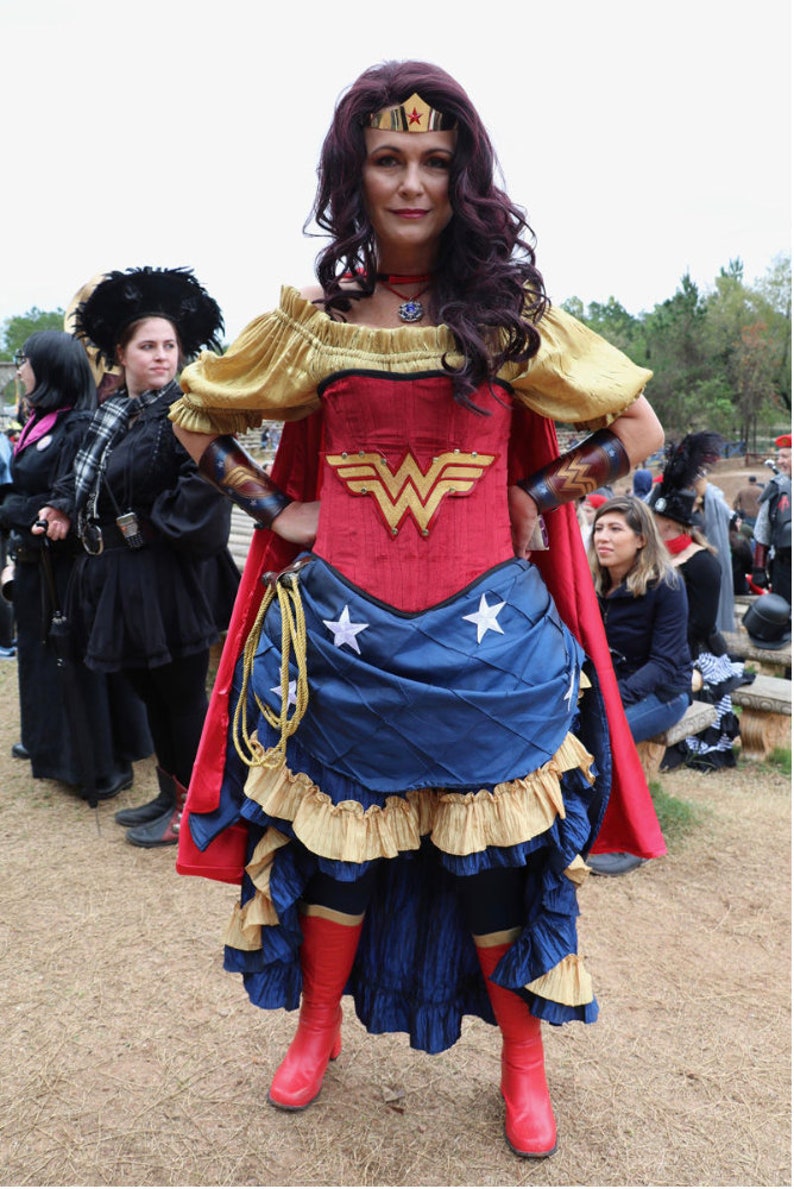 Puede incluir: Una mujer vestida como Wonder Woman con un traje rojo y azul con una falda de estrellas doradas y una capa roja. Lleva una corona dorada con una estrella roja en la parte superior. Est&aacute; de pie con las manos en las caderas.