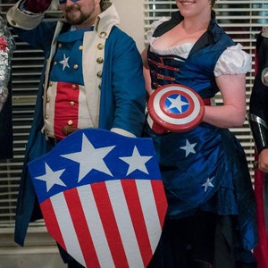 May include: Two people dressed in red, white and blue costumes. The person on the left is wearing a blue jacket, a white shirt with stars, and a red waistcoat. The person on the right is wearing a blue dress with white trim and a red belt. Both people are holding a red, white and blue shield with stars.