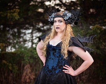 May include: A woman wearing a black dress with a blue bodice and a black and silver floral crown. She has black wings with a spiderweb design.