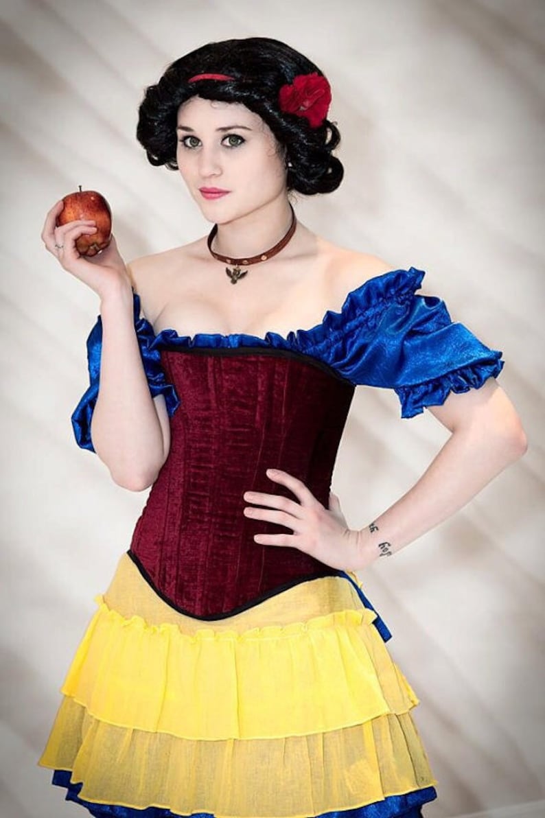 May include: A woman dressed as Snow White, wearing a red and black corset, a blue and white ruffled skirt, and a red flower in her hair. She is holding a red apple in her hand.